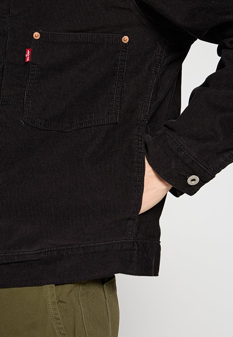 La chaqueta de pana negra cuenta con un bolsillo en el pecho, botones de cobre y puños abotonados. Tejido texturizado con detalle de estrías horizontales.