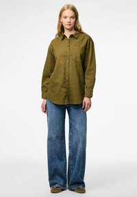 Olivgrön långärmad skjorta med knappar och en frontficka, ihopparad med löst sittande blå jeans och bruna skor.