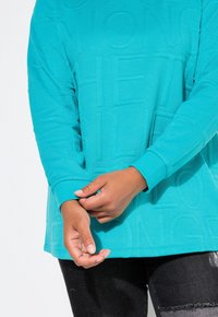 Felpa color teal con design in rilievo, maniche lunghe e polsini a costine. Abbinata a jeans in denim scuro. Texture liscia evidente.