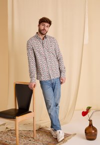 Button-up shirt met een bloemmotief in roze, blauw en groen, gecombineerd met lichtblauwe jeans en witte sneakers, leunend op een stoel.