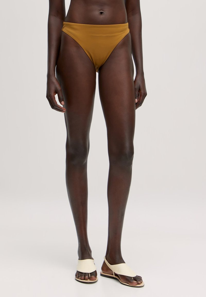 Brauner Bikiniunterteil mit einem hochgeschnittenen Design, glatter Textur und minimalistischem Stil. Kombiniert mit cremefarbenen flachen Sandalen mit einem gespaltenen Riemen.
