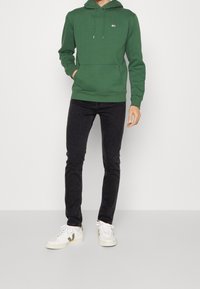 Sudadera verde con capucha y bolsillo delantero, combinada con jeans negros de corte ajustado y zapatillas blancas con un logo dorado.
