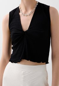 Schwarzes Crop-Top aus strukturiertem Stoff, mit tiefem V-Ausschnitt, gedrehtem Frontdetail und gewelltem Saum, kombiniert mit weißen hochgeschnittenen Hosen.