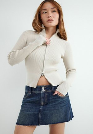 Młoda kobieta ubrana w beżowy sweter zapinany na zamek ze ściągaczami oraz ciemnoniebieską jeansową mini spódniczkę z guzikami, pozująca z jedną ręką na biodrze.