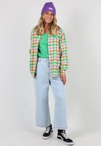 Chemise à carreaux multicolores avec un t-shirt vert en dessous, jean large bleu clair, et baskets noires montantes. Bonnet en tricot violet sur la tête.