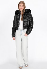 Błyszcząca czarna kurtka puchowa z kapturem wykończonym futrem, zapinana na zamek błyskawiczny, z ściągaczami przy mankietach, noszona na białym topie i zestawiona z jasnymi dżinsami.