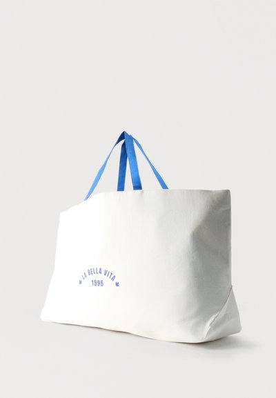 Λευκή τσάντα καμβά με μπλε λαβές και την επιγραφή «LA BELLA VITA 1995» σε μπλε στο μπροστινό μέρος, όρθια σε ανοιχτό φόντο.