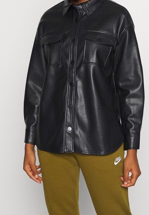 Person wearing a black, buttoned, long-sleeve faux leather shirt with two chest pockets and olive Nike sweatpants.