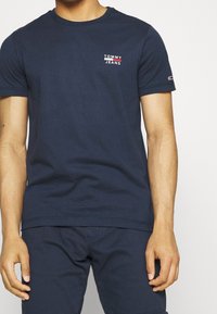 Marinblå bomullströja med rund halsringning, korta ärmar och en liten vit och röd "TOMMY JEANS"-logotyp på vänster bröstområde.