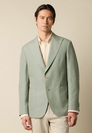 Man draagt lichtgroene blazer over een licht gestreept overhemd en beige broek, staand tegen een beige achtergrond.