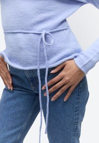 Personne portant un pull tricoté bleu clair avec une ceinture à la taille, associé à un jean en denim bleu, les mains posées sur les hanches.