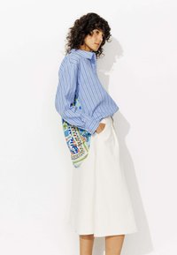 Chemise rayée bleu et blanc à manches longues, associée à une jupe midi blanche. Présente un tissu à motifs colorés drapé sur l'épaule.