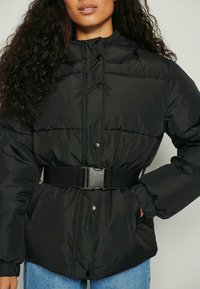 Mujer vestida con una chaqueta acolchada negra con botones a presión, cinturón ajustable, puños el�ásticos y jeans azules, posando con una mano en el bolsillo.