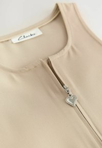 Top senza maniche beige con scollatura rotonda e zip frontale dotata di un tirante a forma di cuore argentato. Texture liscia a coste.