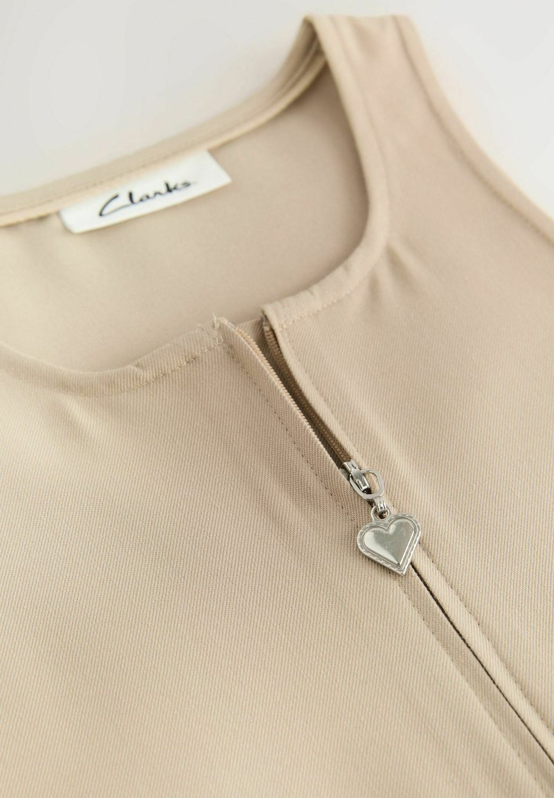 Top senza maniche beige con scollatura rotonda e zip frontale dotata di un tirante a forma di cuore argentato. Texture liscia a coste.