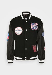 Czarna kurtka varsity z haftowanymi naszywkami, z grafiką baseballową i numerem "85" na piersi. Satynowe rękawy z prążkowanymi mankietami.