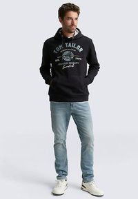 Schwarzer Kapuzenpullover mit Frontmotivdruck, aus Baumwollmaterial. Hellblaue Jeans und weiße High-Top-Sneaker vervollständigen das Outfit.