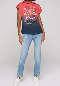 Camiseta con capucha en degradado rojo y marino con texto impreso en blanco, combinada con vaqueros de mezclilla azul claro con detalle de texto lateral y zapatillas blancas.