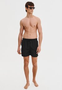 Czarne szorty kąpielowe z elastycznym paskiem i sznurkiem do regulacji. Model nosi ciemne okulary przeciwsłoneczne i stoi boso na jednolitym tle.