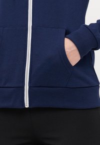 Navy zip-up hoodie with white zipper, ribbed cuffs, and kangaroo pockets. Made from soft material with a smooth texture.