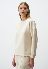 Felpa beige a girocollo con maniche lunghe, vestibilità comoda, texture morbida e polsini a coste, abbinata a pantaloni chiari coordinati.
