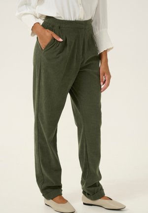 Vrouw draagt olijfgroene ribfluwelen broek met elastische tailleband en beige platte schoenen, met hand in de zak, staand tegen een lichte achtergrond.