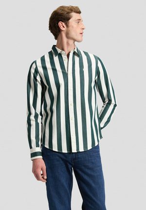 Mann trägt ein langärmeliges Hemd mit vertikalen grünen und weißen Streifen und blaue Jeans, schaut nach rechts und hat eine Hand hinter dem Rücken.