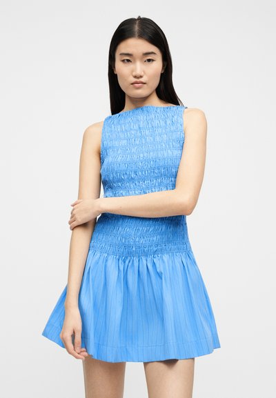 Νεαρή γυναίκα με ίσια σκούρα μαλλιά φοράει αμάνικο, γαλάζιο φόρεμα με σφηκοφωλιά, στέκεται με σταυρωμένα τα χέρια μπροστά σε απλό φόντο.