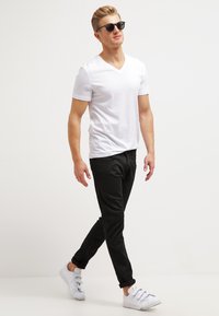 White short-sleeve v-neck t-shirt, black slim-fit pants, and white sneakers. Model wearing black sunglasses, walking on a neutral background.