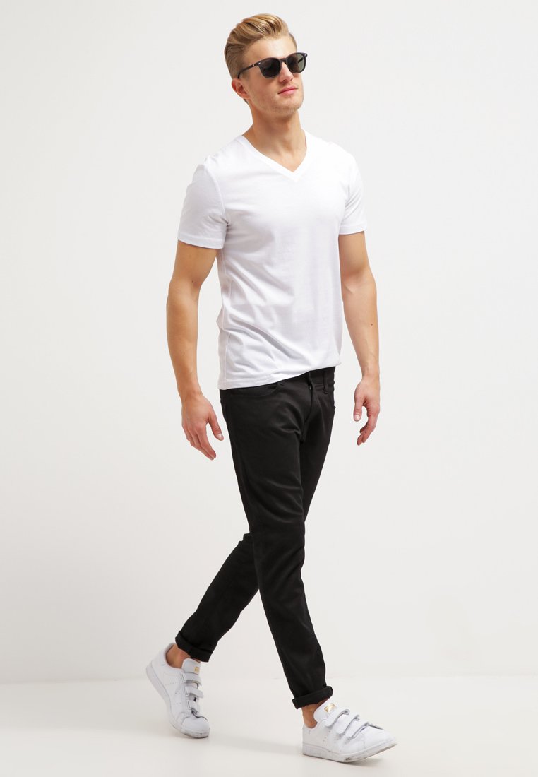 White short-sleeve v-neck t-shirt, black slim-fit pants, and white sneakers. Model wearing black sunglasses, walking on a neutral background.