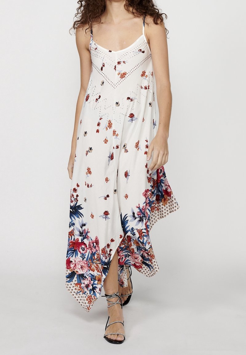 Femme portant une robe longue blanche à fleurs avec des bretelles spaghetti et un ourlet en pointe, associée à des sandales à talons hauts noires à lanières.