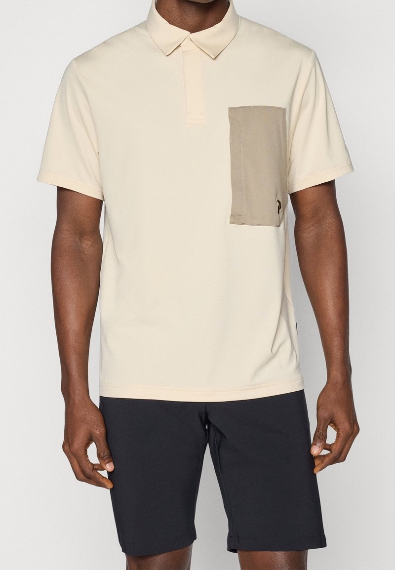 Polo shirt beige avec un col, des manches courtes et une poche poitrine beige contrastante. Porté avec un short noir, affichant une coupe décontractée.