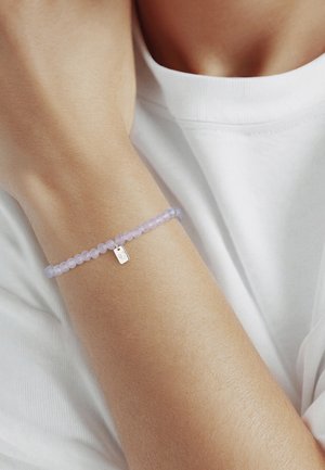 Poignet portant un délicat bracelet lavande à perles avec un petit charme en argent, sur fond de haut blanc sans manches.