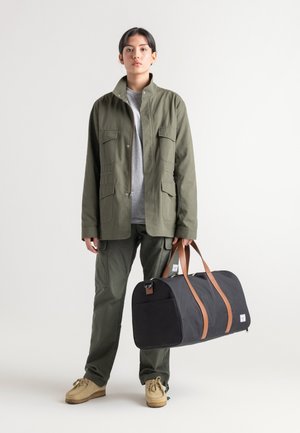 Veste utilitaire en coton vert avec plusieurs poches, t-shirt gris, pantalons cargo verts et un sac de sport noir avec des poignées marron.