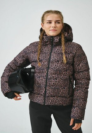 Jeune femme aux cheveux tressés porte une doudoune à imprimé léopard et tient un casque de ski noir, debout devant un fond uni.