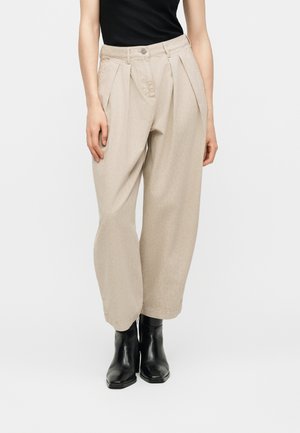 Γυναίκα με μπεζ ψηλόμεσο πλισέ παντελόνι, μαύρο αμάνικο τοπ και μαύρα μποτάκια, στέκεται μπροστά σε απλό φόντο.