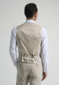Chaleco formal beige con un acabado suave, correa trasera ajustable y una textura ligera, acompañado de una camisa blanca de manga larga.