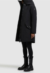 Personne portant un long manteau d'hiver noir avec capuche, des leggings noirs et des bottines noires sur un fond gris clair uni.