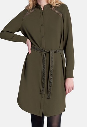 Femme portant une robe boutonnée à manches longues vert olive avec détail en dentelle sur les épaules et une ceinture assortie en dentelle nouée à la taille.