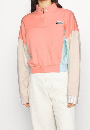 Kobieta w pastelowej, kolorowej bluzie Adidas z panelami w kolorze koralowym, beżowym i jasnoniebieskim, w połączeniu z białymi, wysokim stanem spodniami.