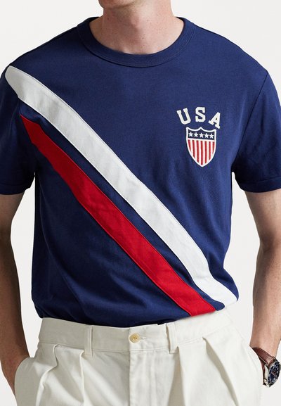 Tricou navy blue cu dungi diagonale albe și roșii, având inscripția "USA" și un emblema în formă de scut pe piept. Material din bumbac cu guler rotund.