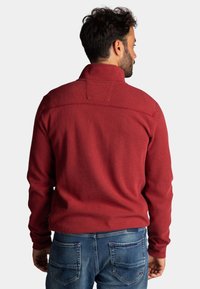 Rode, textuurtrui met een hoge kraag, voorzien van een gestikte baan over de schouders. Draagt met blauwe denim jeans.