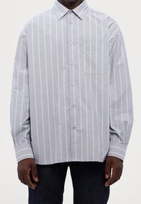 Camisa de manga larga azul claro con rayas verticales blancas, cuello abotonado, un bolsillo en el pecho y un corte relajado. Material de algodón suave.