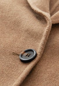 Tissu de manteau en laine beige avec une texture lisse, doté d'un grand bouton noir à deux trous et d'un détail de couture subtil.