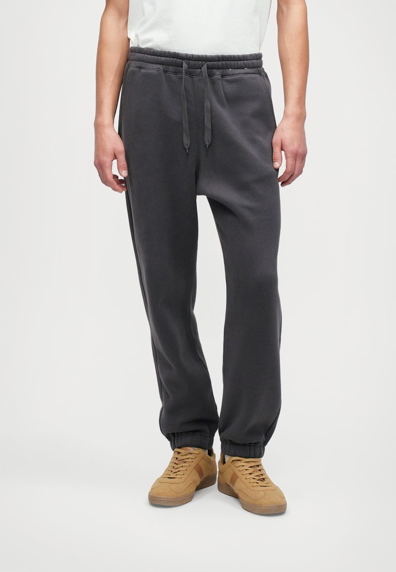 Pelēki kokvilnas sporta bikses ar elastīgu jostasvietu, regulējamu auklu un pagarinātiem svārkiem, savienotas ar brūnām sporta apaviem.