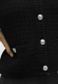 Zwarte gebreide cardigan met een textuurpatroon, geribbelde randen en zilverkleurige knopen, voorzien van twee voorke pockets aan de onderzijde.