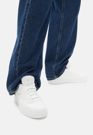 Białe skórzane trampki z perforowanym przodem, płaskimi sznurowadłami i wyściełanym językiem, noszone z luźnymi, ciemnoniebieskimi dżinsami.