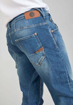 Jean en denim bleu avec une coupe classique, présentant une étiquette en cuir marron à la taille et un design de poche angulaire distinctif sur la cuisse.