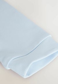 Heller blauer, gerippter Stoffbund eines Kleidungsstücks mit glatter Textur und abgerundeter Kante, vor einem neutralen Hintergrund präsentiert.