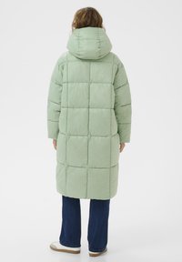 Osoba ubrana w długi, jasnoszary pikowany płaszcz z kapturem, ciemnoniebieskie dżinsy i białe trampki, stoi tyłem na białym tle.